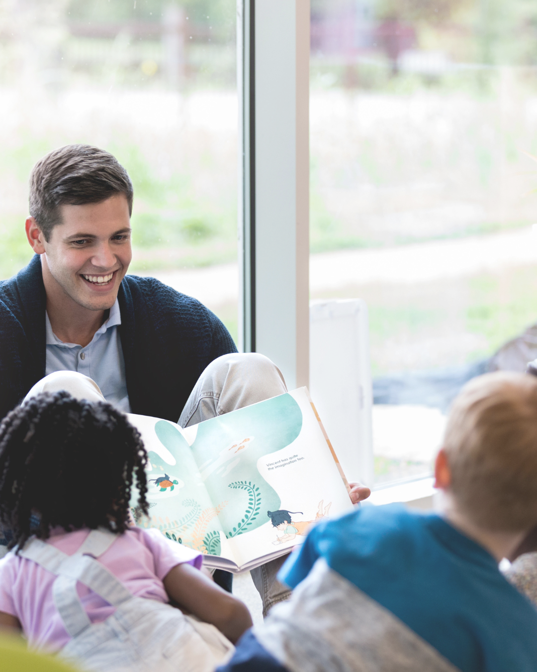 Pädagoge liest Kindern im Kindergarten aus einem Bilderbuch vor – ideale Umgebung für sensorisches Spiel mit Therapieschaukel